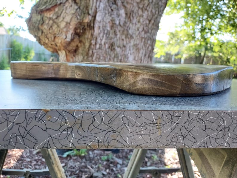 Op de afbeelding: Een houten snijplank in donkerbruin met een handvat. De plank heeft een gladde, gebogen vorm en ligt op een grijze ondergrond met een wit en zwart wervelpatroon. Op de achtergrond zijn een boom en groen te zien.