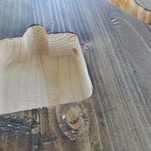 Op de afbeelding: Close-up van een houten oppervlak met een uitgesneden gedeelte. Het hout is grijs gebeitst en het uitgesneden gedeelte is van natuurlijk hout. Het ontwerp suggereert een handvat of een verzonken kenmerk.