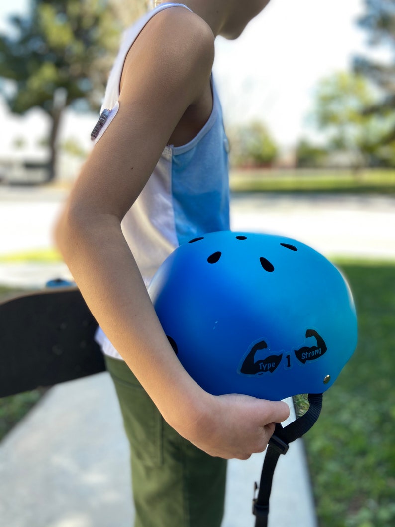 May include: A blue helmet with black stickers that say "Type 1 Strong" on the side. The helmet is being held by a person.