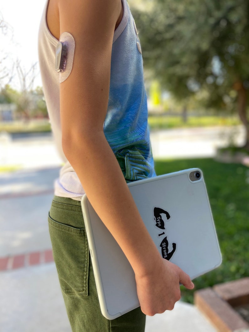 May include: A young person wearing a blue and white tank top with a medical device on their arm, holding a white tablet with a black sticker that says "Type 1 Strong".