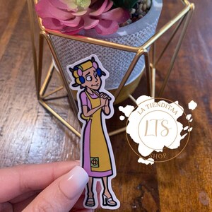 May include: A yellow and pink cartoon sticker of a woman with a yellow apron and blue hair rollers. The sticker has a small flower design on the apron and the text "La Tiendita LTS Shop" is on a white circle behind the woman.