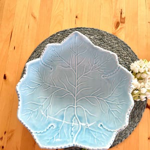 Pode incluir: Uma travessa de cerâmica azul clara em forma de folha, com padrões de veias detalhados. A travessa está sobre um tapete cinza tecido, com flores brancas e caules verdes no fundo. Uma peça decorativa para a casa.