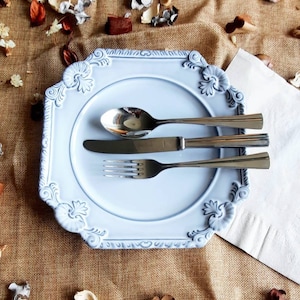 Puede incluir: Un plato azul claro adornado con una cuchara, un cuchillo y un tenedor. El plato tiene un diseño floral decorativo en los bordes. La cubertería es de acero inoxidable. El conjunto está sobre una superficie de arpillera.