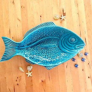 以下が含まれることがあります： 光沢のあるターコイズブルーの魚の形をした陶器製の盛り付け皿。鱗、鰭、目が細かく浮き彫りにされています。皿の周りには、小さな装飾用の貝殻、白いヒトデ、滑らかな青いガラスの小石が配置されています。