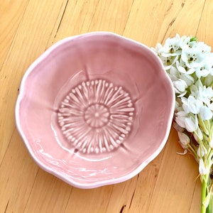 May include: A light pink ceramic bowl with a flower-like design. The bowl has a textured center and a wavy rim. White flowers with green stems are placed to the right of the bowl. The bowl is on a light brown wooden surface.