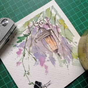 Puede incluir: Pintura acuarela de una farola con un fondo verde, morado y rosa. La farola está colgada de una rama y tiene un acabado dorado. La pintura está firmada con "2021".