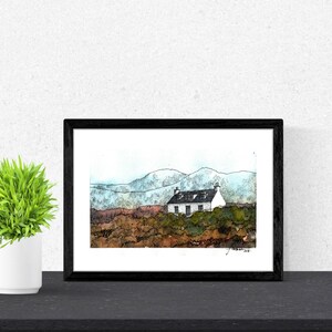 May include: A framed watercolor painting of a white cottage with a black roof, set against a backdrop of blue and green mountains. The cottage is surrounded by brown and green brush.