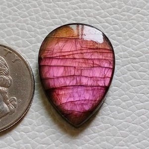 May include: A teardrop-shaped cabochon gemstone displaying a vibrant pink and magenta surface with horizontal crackled textures and a smooth, polished finish, positioned next to a circular metal coin for scale.