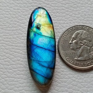 Può includere: Una gemma di labradorite ovale con colori iridescenti blu, verdi e gialli. Accanto, una moneta da un quarto di dollaro USA (2,4 cm di diametro) per il confronto delle dimensioni. La superficie lucida riflette la luce, mostrando i suoi motivi naturali.