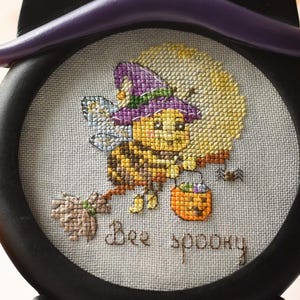 May include: Black, cat-shaped Halloween decoration with a witch hat. The circular center features a cross-stitch design of a bee witch with the text "Bee Spooky". The decoration sits on a stack of books, with a candle in the background.