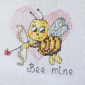 Puede incluir: Bordado de punto de cruz en un aro de madera con una abeja de dibujos animados con arco y flecha, sobre un corazón rosa. El texto "Bee mine" está bordado debajo. El fondo incluye flores amarillas secas y partituras.