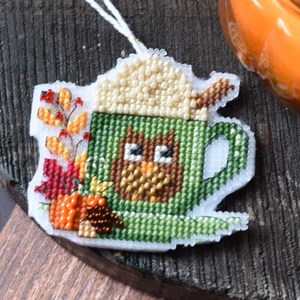 Può includere: Un ornamento a punto croce a forma di tazza di caffè con foglie autunnali e una zucca. La tazza è verde con un disegno di caffè marrone e panna montata color crema. Una candela arancione accesa e un vaso verde a forma di stivale sono sullo sfondo.