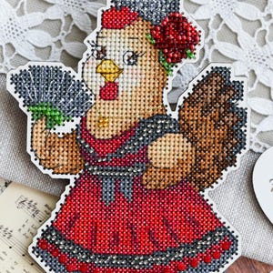 Könnte beinhalten: Ein Kreuzstich-Design einer Henne in einem roten Kleid, die einen Fächer hält und eine Blume im Haar trägt. Das Bild enthält einen Leinenbeutel mit Spitzensaum, Noten und ein Etikett mit der Aufschrift "From Spain with Love".