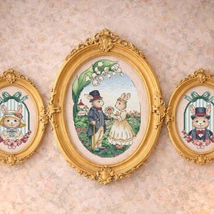 Könnte beinhalten: Drei gerahmte Kreuzstichbilder mit goldenen, verzierten Rahmen, die Kaninchen in viktorianischer Kleidung zeigen. Darunter ein weißer Kaminsims mit Blumenarrangements, Kerzen und Büchern. Die Szene wirkt sanft und romantisch.