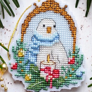Könnte beinhalten: Ein Kreuzstich-Weihnachtsschmuck mit einem weißen Vogel mit blauem Schal, goldfarben gerahmt. Der Vogel ist von einem Kranz mit einer Kerze und roten Schleifen umgeben. Der Schmuck liegt vor Tannenzweigen und goldenen Perlen.
