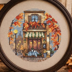 Könnte beinhalten: Eine kreisförmige Kreuzstichstickerei in einem dunklen Holzrahmen, die ein mehrstöckiges Gebäude mit Balkon, herbstlichem Laub und ein Straßencafé darstellt. Das Design zeigt warme Orange-, Gelb- und Brauntöne auf hellem Stoff.