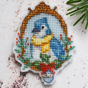 May include: A cross-stitched Christmas ornament featuring a blue bird wearing a yellow scarf and a hat, framed in a gold-colored border. The ornament is decorated with red berries and green foliage, with a small red bow at the bottom.