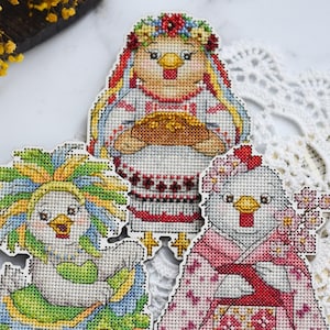 Könnte beinhalten: Drei Kreuzstich-Hühnerfiguren, jede in einem einzigartigen Outfit. Eine trägt einen Blumenkranz und ein besticktes Kleid, eine andere ein buntes Rüschenkleid und die dritte einen rosa Kimono mit floralen Akzenten. Die Figuren befinden sich auf einer weißen Oberfläche.