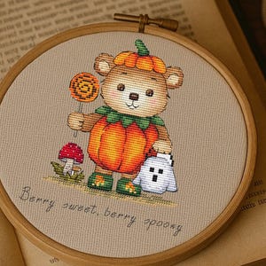 Könnte beinhalten: Kreuzstich-Stickerei in einem Holzrahmen mit einem Bären im Kürbiskostüm, der einen Lolli und einen Geist hält. Der Text "Berry sweet, berry spooky" ist darunter gestickt. Eine brennende Kerze, ein Tintenfass und ein offenes Buch sind im Hintergrund.