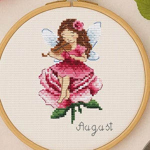 Könnte beinhalten: Ein Kreuzstich-Stickrahmen mit einer Fee auf einer Rose, umgeben von rosa Rosen, Garn und einer goldenen Schere. Das Wort "August" ist darunter gestickt. Die Farbpalette umfasst Rosa, Grün und Beige.