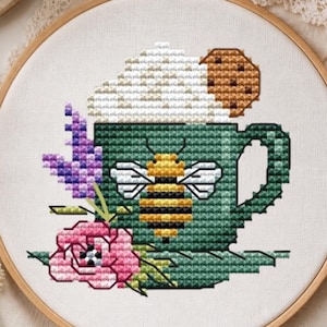 Könnte beinhalten: Eine Kreuzstichstickerei in einem Holzrahmen zeigt eine grüne Tasse mit Bienenmotiv, gekrönt von weißem Schaum und einem braunen Keks. Die Tasse ist mit einer rosa Blume und violetten Zweigen auf weißem Stoffgrund verziert.