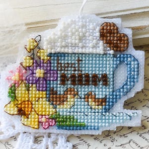 Könnte beinhalten: Ein kreuzgestickter Anhänger in Form einer blauen Tasse mit dem Wort „MUM“. Die Tasse ist mit Blumen und Herzen verziert. Der Anhänger liegt auf einer weißen Spitzendecke.