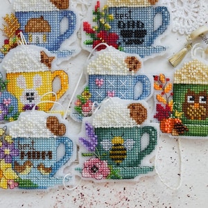 Könnte beinhalten: Eine Sammlung von sieben Kreuzstich-Anhängern in Tassenform mit verschiedenen Motiven wie einer Biene, einer Eule und den Schriftzügen best dad und best mom. Jede Tasse ist mit weißem Schaum und einem kleinen Keks sowie bunten Blumen verziert.