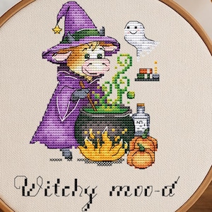 Könnte beinhalten: Ein Kreuzstich-Stickrahmen mit einer Kuh im Hexenkostüm, die einen Kessel umrührt. Das Design umfasst einen Geist, Kürbisse und den Text "Witchy moo-d". Der Rahmen wird mit Kürbissen und einer brennenden Kerze präsentiert.