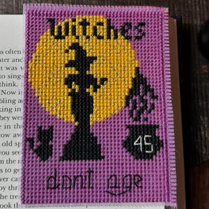 Könnte beinhalten: Ein lila Kreuzstich-Lesezeichen mit dem Wort "Witches" und einer Hexensilhouette vor einem gelben Mond. Das Lesezeichen hat auch die Worte "don't age" und die Zahl 45.