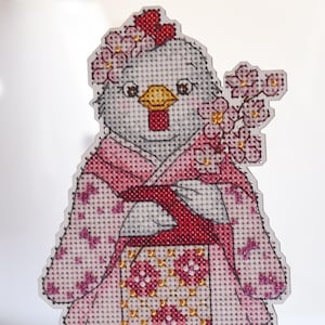 Könnte beinhalten: Eine Kreuzstich-Figur eines Huhns in einem rosa Kimono mit floralen Mustern und einer roten Schleife. Das Huhn hält einen Zweig mit rosa Blüten. Die Figur steht auf einem kleinen Holzsockel. Der Hintergrund ist verschwommen, mit gelben Blumen in einer weißen Vase.
