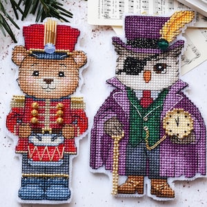 May include: Two cross-stitched Christmas ornaments: a bear dressed as a toy soldier and an owl in a purple coat and top hat. The bear wears a red uniform and blue pants. The owl has a monocle and holds a pocket watch.
