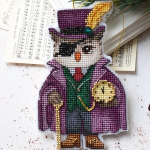 Könnte beinhalten: Ein Kreuzstich-Eulen-Ornament mit lila Zylinder, Mantel und Augenklappe, mit Gehstock und Taschenuhr. Die Eule hat goldene und grüne Akzente. Der Hintergrund zeigt Notenblätter, eine Kerze und grünes Blattwerk.