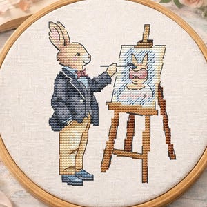 Può includere: Un telaio da ricamo a punto croce con un coniglio artista che dipinge un autoritratto. Il coniglio indossa una giacca blu e pantaloni beige. La scena comprende un libro, fiori e una tazza di tè.