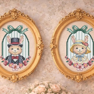 Könnte beinhalten: Zwei gerahmte Kreuzstichporträts von Hasen, einem Männchen und einem Weibchen, in verzierten ovalen Goldrahmen. Das männliche Kaninchen trägt einen Zylinder und einen Smoking, während das weibliche Kaninchen eine Haube trägt. Beide sind von floralen Akzenten umgeben.