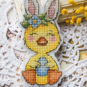 Könnte beinhalten: Ein Kreuzstich-Osteranhänger in Form eines gelben Küken mit Hasenohren. Das Küken hält ein blaues Ei und trägt einen Blumenkranz. Der Anhänger hat eine weiße Schnur zum Aufhängen und liegt vor Spitze und Trockenblumen.