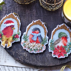 Könnte beinhalten: Drei gestickte Weihnachtsschmuckstücke mit roten Vögeln in verzierten Rahmen. Ein Vogel trägt einen Weihnachtsmannhut, ein anderer einen blauen Schal und der dritte einen grünen Hut. Goldband und Kerze im Hintergrund.