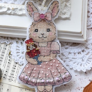 Könnte beinhalten: Ein Kreuzstich-Hasenornament in einem rosa Kleid, das einen Spielzeugsoldaten hält. Der Hase hat eine rosa Schleife und hängt an einer Schnur. Eine brennende Kerze in einem Glas steht im Hintergrund, zusammen mit Noten und anderen kleinen Gegenständen.