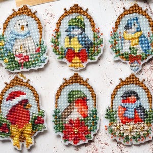 May include: Six embroidered bird ornaments with gold-colored frames. Each features a different bird design, including a cardinal in a Santa hat, a bluebird with a scarf, and a robin with a poinsettia. The ornaments are surrounded by festive greenery.