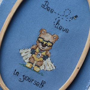 Könnte beinhalten: Kreuzstich-Design auf blauem Stoff, das einen Bären-Superhelden mit Brille und Umhang zeigt. Der Text "Bee-lieve in yourself" ist in schwarzem Faden gestickt. Das Design befindet sich in einem hölzernen Stickrahmen.