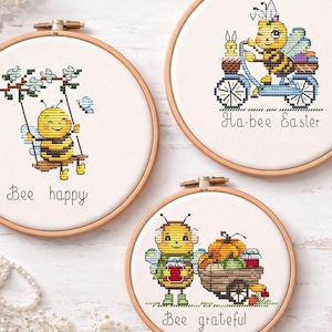 Könnte beinhalten: Drei Kreuzstichreifen mit Bienenmotiven. Einer zeigt eine Biene auf einer Schaukel mit dem Text "Bee happy". Ein anderer zeigt eine Biene auf einem Fahrrad mit "Ha-bee Easter". Der letzte zeigt eine Biene mit einem Wagen voller Kürbisse und dem Text "Bee grateful".