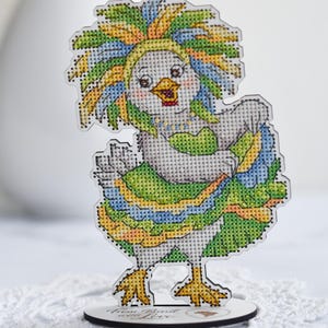 Könnte beinhalten: Ein kreuzgesticktes Hühnchen-Figürchen mit buntem Kopfschmuck und Rüschenrock. Das Hühnchen hat einen gelben Schnabel und steht auf einem Holzsockel mit Text. Die Figur ist etwa 10 cm hoch.