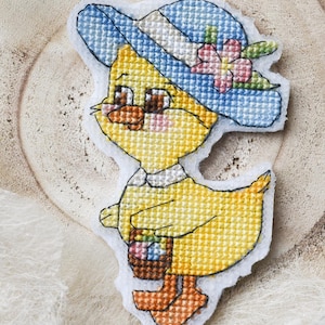 Könnte beinhalten: Ein kreuzgesticktes gelbes Küken mit blauem Hut und rosa Blume, das einen Korb trägt. Das Küken hat braune Augen und orangefarbene Füße. Das Kunsthandwerk befindet sich auf einer hellen, strukturierten Oberfläche, mit einem weißen Faden befestigt.