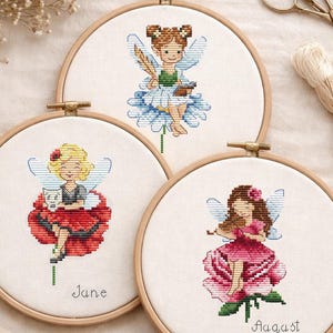 Könnte beinhalten: Drei Kreuzstich-Stickrahmen mit Feen-Designs. Jede Fee wird in einer anderen Pose dargestellt, die auf einer Blume sitzt. Die Reifen sind vor einem neutralen Hintergrund mit Bastelzubehör wie Schere und Faden angeordnet.