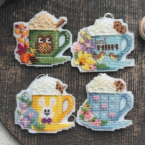 Könnte beinhalten: Vier Kreuzstich-Ornamente in Form von Kaffeetassen, jedes mit einem anderen Design. Eines ist grün mit einer Eule, ein anderes blau mit dem Wort "MUM", ein gelbes mit einem Hasen und ein blaues mit Blumen. Alle haben Schlagsahne.