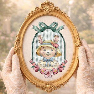 Puede incluir: Un punto de cruz ovalado con marco dorado de un conejito con sombrero y vestido, rodeado de flores rosas y un lazo verde. El marco está sostenido por manos con guantes de encaje, sobre un fondo de jardín borroso.