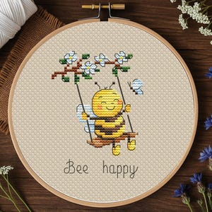 Könnte beinhalten: Kreuzstich-Stickerei in einem Holzrahmen zeigt eine lächelnde Biene auf einer Schaukel, mit dem Text "Bee happy" darunter. Die Biene ist gelb und schwarz, schwingt an einem Ast mit weißen Blüten. Eine Spule weißen Garns und Blumen sind im Hintergrund.