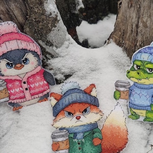 Könnte beinhalten: Drei Kreuzstichfiguren von Tieren in Winterkleidung, die Kaffeetassen halten, in einer verschneiten Szene. Ein Pinguin mit rosa Hut und Mantel, ein Fuchs mit blauem Hut und grünem Mantel und ein Alligator mit blauem Pullover.