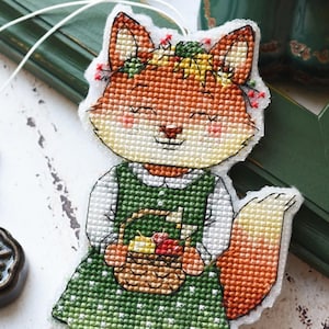 Könnte beinhalten: Ein Kreuzstich-Ornament, das einen Fuchscharakter in einem grünen Kleid mit weißen Punkten, einem weißen Hemd und gelben Schuhen zeigt. Der Fuchs trägt einen Blumenkranz, hält einen kleinen Obstkorb und hat einen großen, buschigen orange-weißen Schwanz.