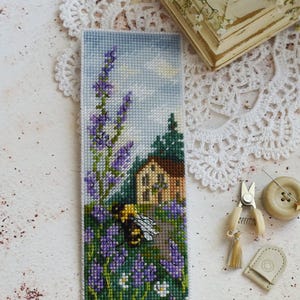Könnte beinhalten: Ein rechteckiges Kreuzstich-Lesezeichen mit einer detaillierten Szene einer Hummel in der Nähe von violetten Lavendelstängeln und weißen Blumen vor einem kleinen Haus mit braunem Dach, vor einem hellblauen Himmel mit weißen Wolken.