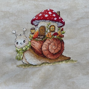 Peut inclure: Une broderie au point de croix sur tissu beige représentant un escargot portant une écharpe vert clair. La coquille de l'escargot est remplacée par une petite maison au toit champignon rouge, porte brune et fenêtres jaunes, entourée de feuilles d'automne.